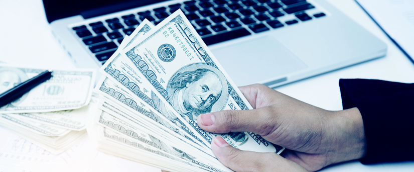 a man’s hands holding dollar banknotes with some more banknotes on the side in front of an open laptop